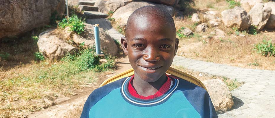 A young Nigerian boy smiles at the camera wearing a blue shirt with his rural village behind him.