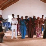 A group of singers with a pianist and a guitarist on either side stood in a building.